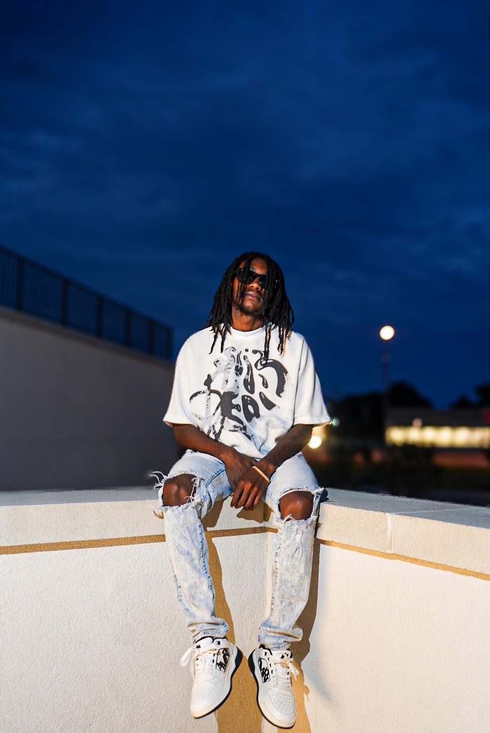 A young man in a graphic tee sitting on a wall at dusk